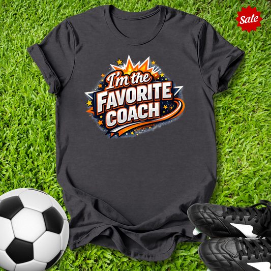Gray t-shirt with 'I'm the Favorite Coach' design on grass with soccer ball and shoes.