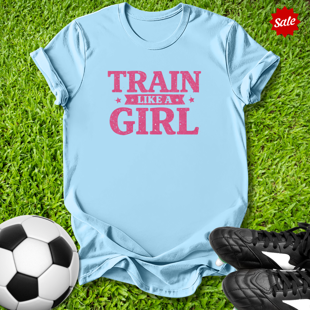 Light blue t-shirt with 'Train Like a Girl' text on grass with soccer ball and shoes.