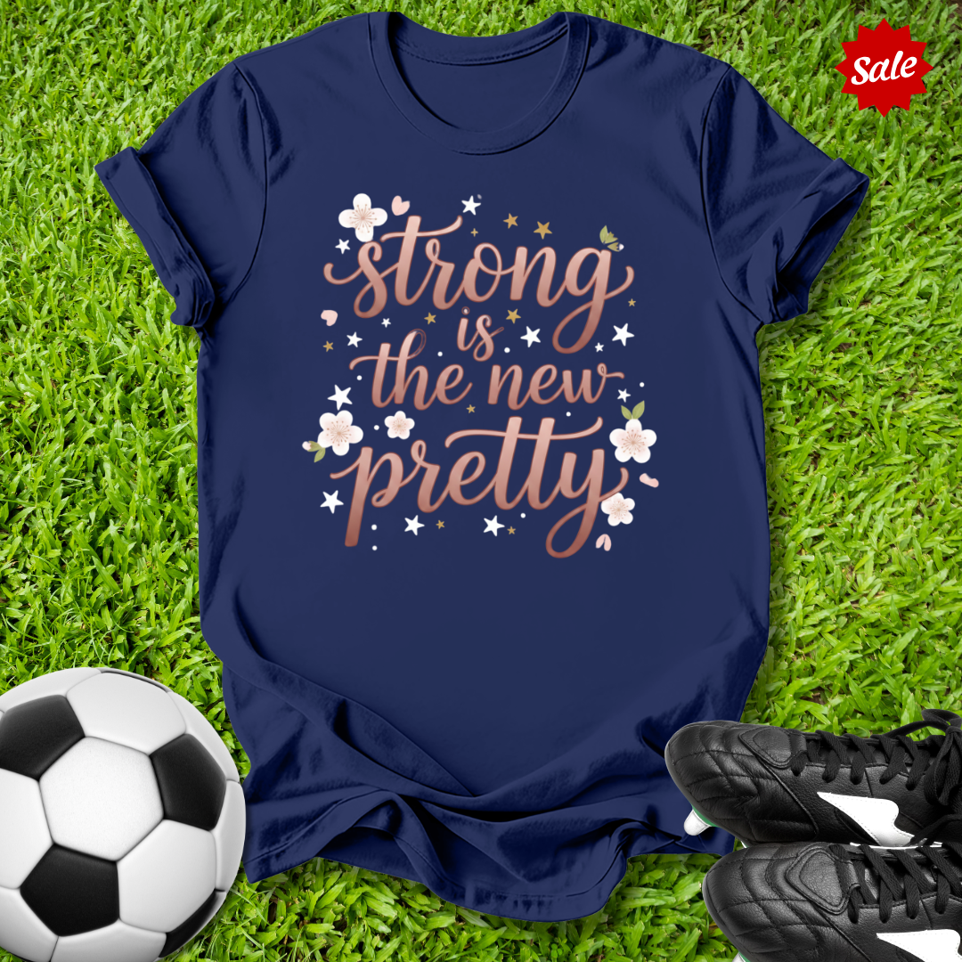 Navy blue t-shirt with 'strong is the new pretty' text on grass with soccer ball and shoes