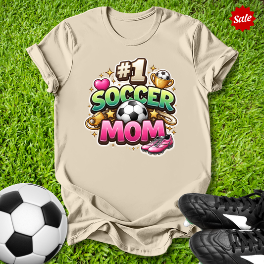 Beige t-shirt with '#1 Soccer Mom' design on grass with soccer ball and shoes.