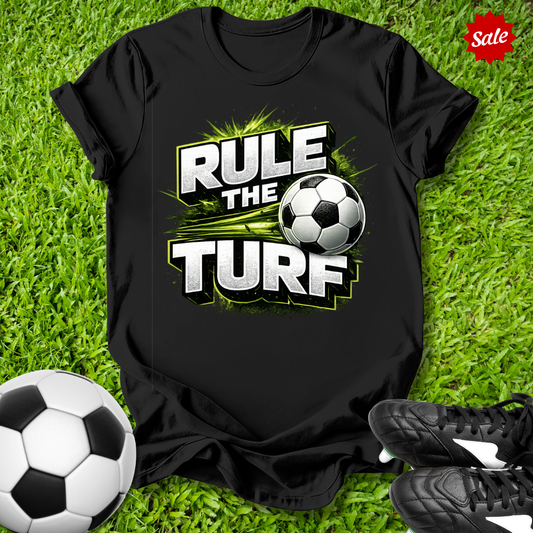 Black t-shirt with 'Rule the Turf' design on grass with soccer ball and shoes.