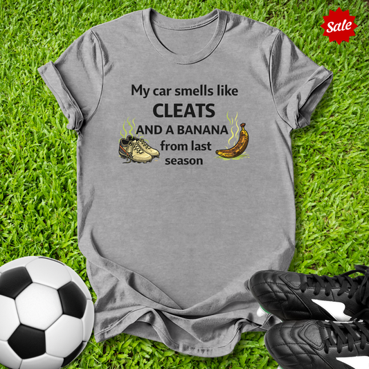 Gray t-shirt with humorous my car smells like cleats and a banana from last season text about car smell, soccer ball, and cleats on grass
