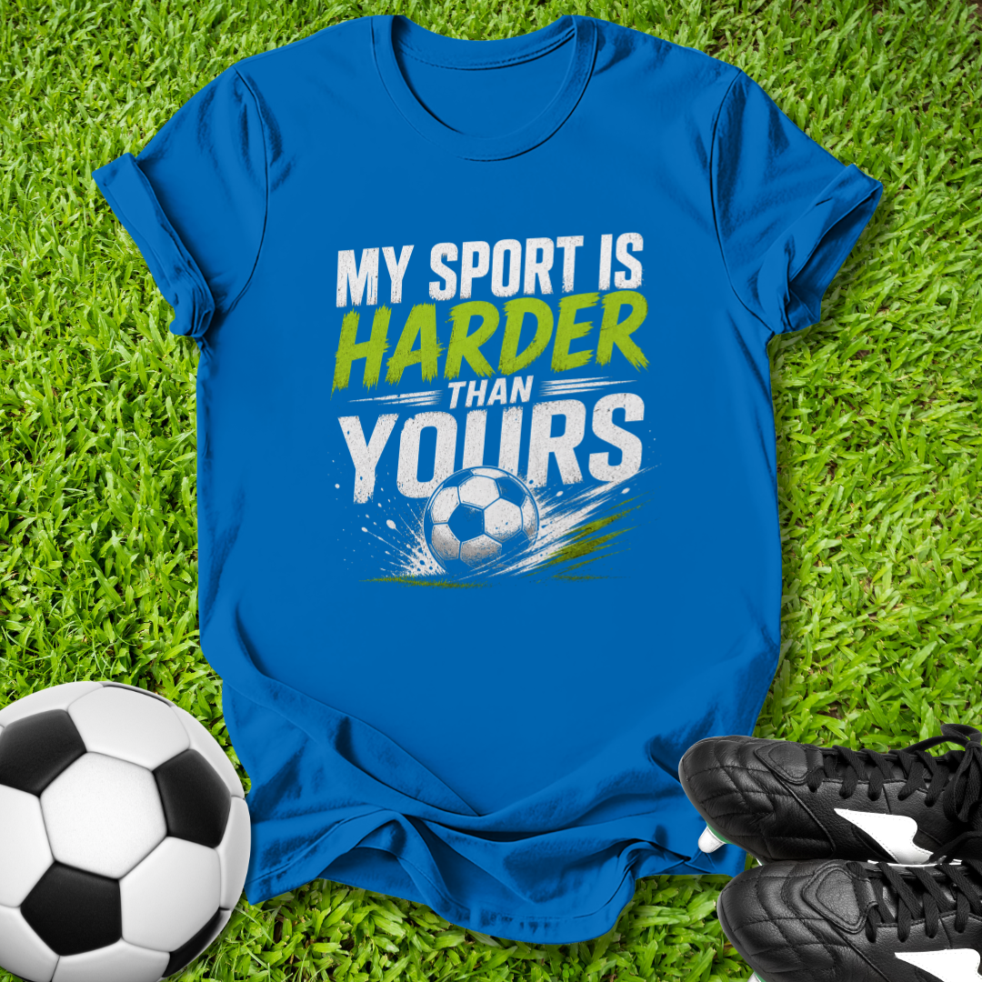 Blue t-shirt with 'My sport is harder than yours' text on grass with soccer ball and shoes.
