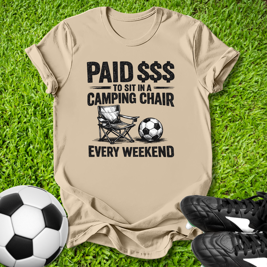 Beige t-shirt with a graphic and text on grass with soccer ball and shoes.