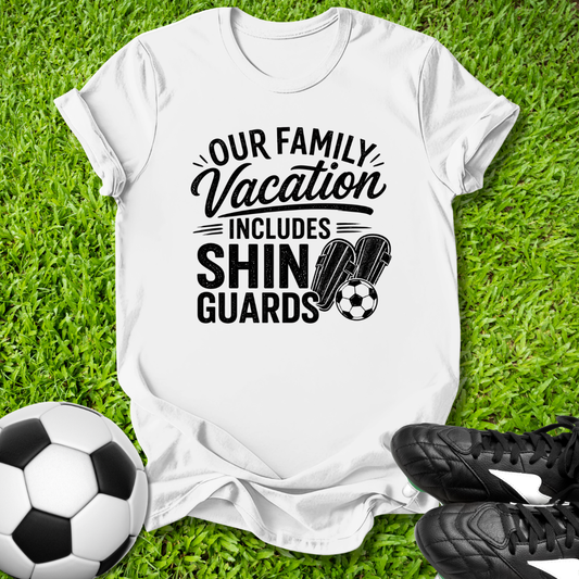 White t-shirt with 'Our Family Vacation Includes Shin Guards' text on grass with soccer ball and shoes.