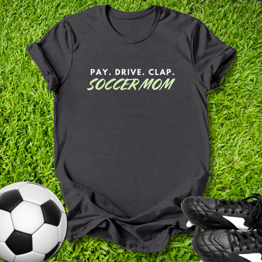 Black t-shirt with 'PAY. DRIVE. CLAP. SOCCER MOM' text on grass with soccer ball and shoes.