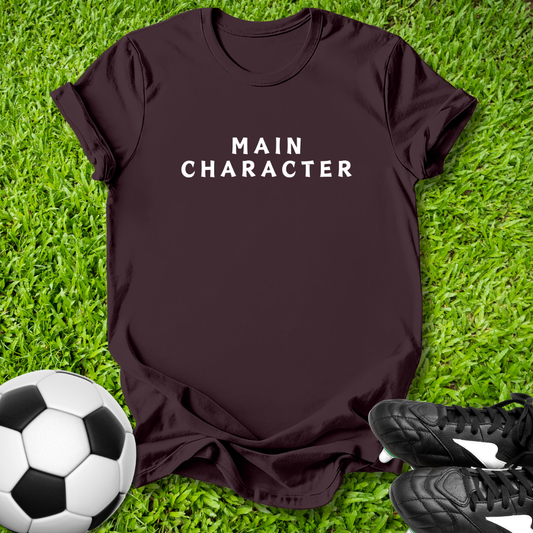 Brown t-shirt with 'MAIN CHARACTER' text on grass with soccer ball and shoes