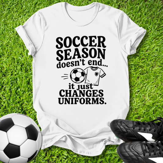 White t-shirt with soccer-themed text on grass with soccer ball and shoes