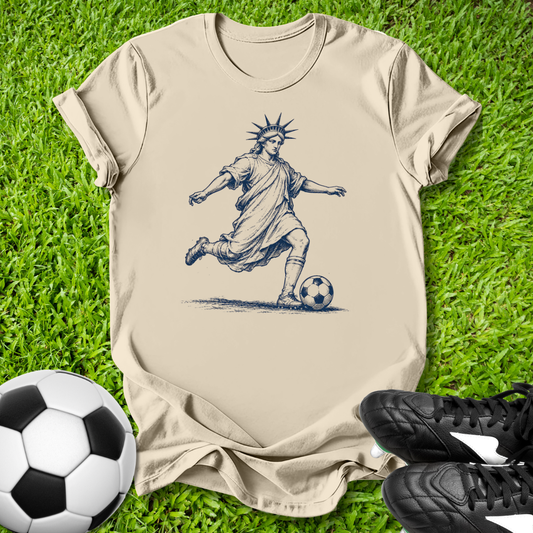 Beige t-shirt with a graphic of a soccer player resembling the Statue of Liberty on grass with soccer balls and shoes.