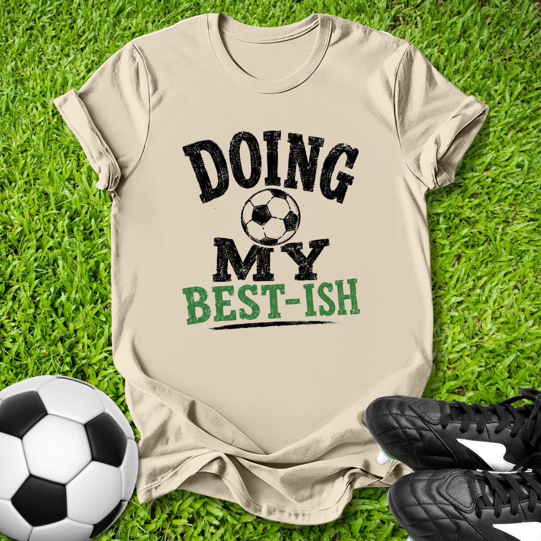 Beige t-shirt with 'Doing my best-ish' text on grass with soccer ball and shoes.