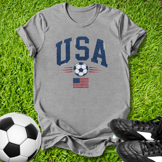 Gray t-shirt with 'USA' and soccer ball design on grass with soccer ball and shoes.