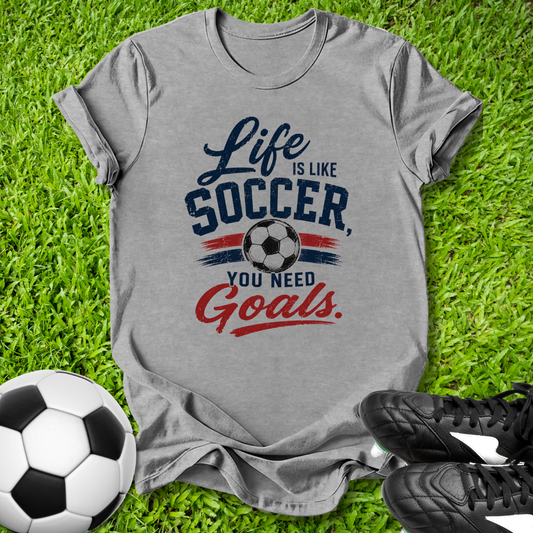 Gray t-shirt with 'Life is like Soccer, you need Goals' text on grass with soccer ball and shoes.
