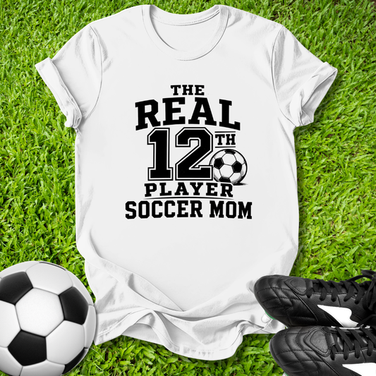 White t-shirt with 'The Real 12th Player Soccer Mom' text on grass with soccer ball and shoes.
