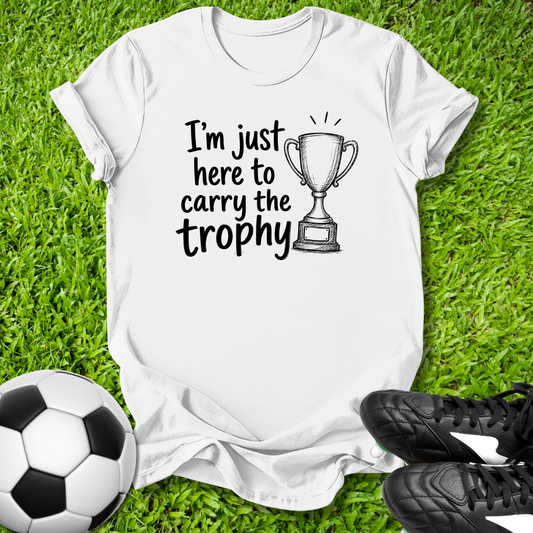 White t-shirt with 'I'm just here to carry the trophy' text and soccer ball and shoes on grass