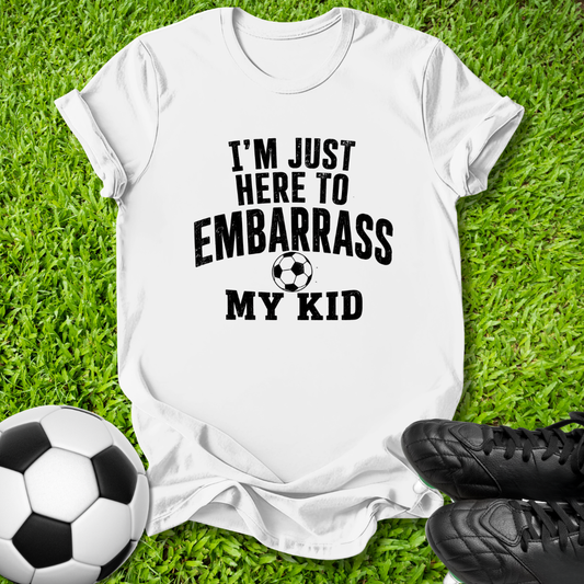 A white short-sleeve graphic T-shirt laid flat on green grass, with black text reading “I'M JUST HERE TO EMBARRASS MY KID” and a soccer ball graphic. A soccer ball and black cleats are placed around it as props.
