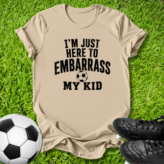 A beige graphic t-shirt laid flat on green grass, featuring the black text 'I'M JUST HERE TO EMBARRASS MY KID' with a soccer ball graphic. A black-and-white soccer ball and black cleats are placed around it as props.