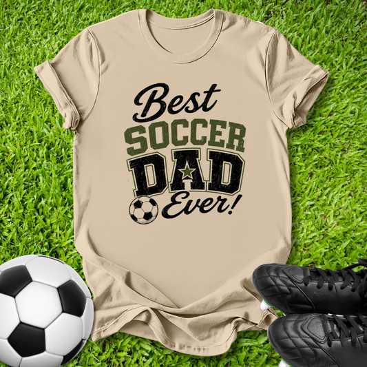 A beige graphic t-shirt laid flat on green grass, featuring the text 'Best SOCCER DAD Ever!' in black and olive green lettering. A soccer ball is on the left and a pair of black cleats is on the right, creating a themed flat-lay composition.