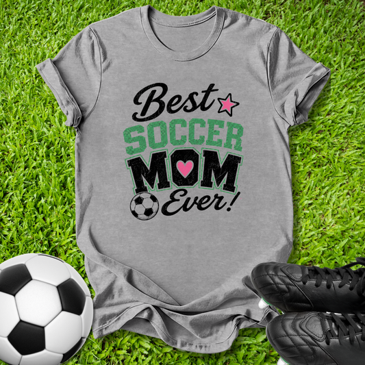 A heather gray graphic t-shirt laid flat on green grass, featuring the text 'Best SOCCER MOM Ever!' with a pink heart and star. A soccer ball is on the left and black cleats are on the right.