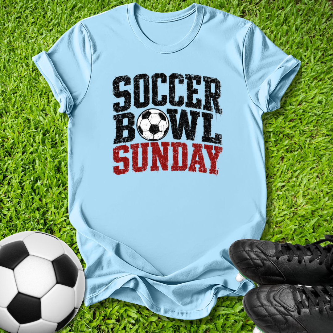 A light blue t-shirt laid flat on green grass, featuring the text 'SOCCER BOWL SUNDAY' in black and red with a soccer ball graphic. A soccer ball and black cleats are placed around it as props.