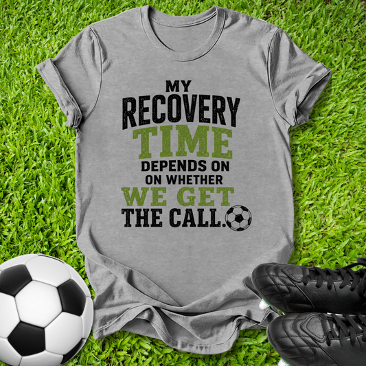 A heather gray graphic t-shirt laid flat on green grass, featuring the text 'MY RECOVERY TIME DEPENDS ON WHETHER WE GET THE CALL.' in black and olive green lettering, accompanied by a soccer ball and black cleats.