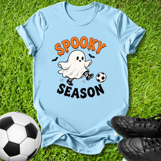 A light blue graphic t-shirt laid flat on green grass, featuring a cartoon ghost kicking a soccer ball with the words 'SPOOKY SEASON' in orange and black. A soccer ball and black cleats are placed around it as props.