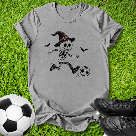 A heather gray short-sleeve t-shirt with a cartoon skeleton soccer graphic, laid flat on green grass with a soccer ball and black cleats as props.