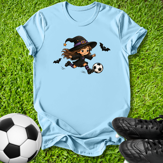 A light blue short-sleeve t-shirt laid flat on green grass, featuring a cartoon graphic of a girl in a witch hat kicking a soccer ball, with two bats flying around. A soccer ball and black cleats are placed around the shirt as props.