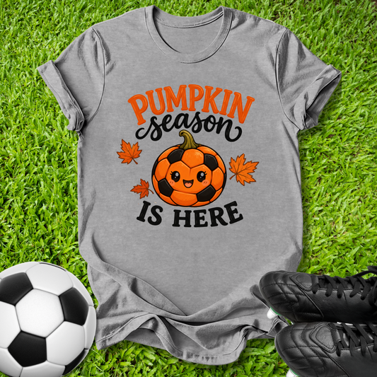 A heather gray graphic t-shirt laid flat on green grass, featuring a cute orange and black soccer pumpkin with the text 'PUMPKIN season IS HERE' and small orange maple leaves. A soccer ball and black cleats are styled around the shirt.
