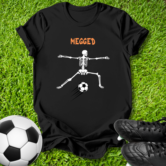 A black short-sleeve graphic T-shirt laid flat on green grass, featuring the word 'MEGGED' in orange above a white skeleton kicking a soccer ball. A soccer ball and black cleats are placed around it as props.