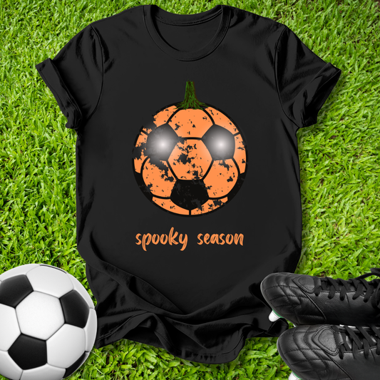 A black short-sleeve T-shirt laid flat on green grass, featuring an orange soccer ball graphic with a pumpkin face and the words 'spooky season' in orange. A black-and-white soccer ball appears at the lower left, and black cleats are at the lower right.