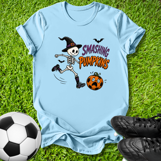 A light blue graphic t-shirt laid flat on green grass, featuring a cartoon skeleton in a witch hat kicking a smiling orange pumpkin with the text 'SMASHING PUMPKINS'. A soccer ball and black cleats are placed around it as props.