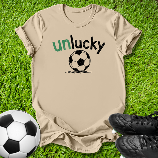 A beige graphic t-shirt laid flat on green grass, featuring the text 'unlucky' in green and black above a black-and-white soccer ball. A soccer ball and black cleats are placed around it as props.