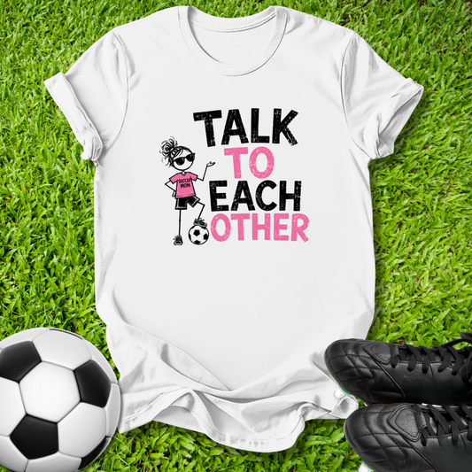 A white graphic t-shirt laid flat on green grass, featuring a cartoon girl in a pink soccer jersey and the text 'TALK TO EACH OTHER' in black and pink. A soccer ball is on the left and black cleats are on the right.