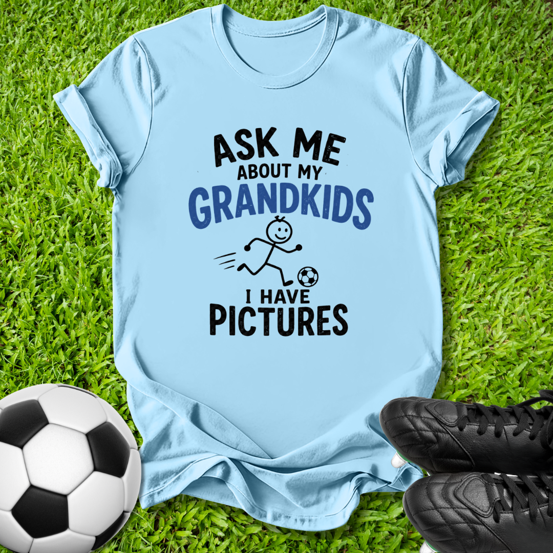 A light blue graphic t-shirt laid flat on green grass, featuring the text 'ASK ME ABOUT MY GRANDKIDS I HAVE PICTURES' in black and blue lettering with a stick figure and soccer ball illustration. A soccer ball and black cleats are placed around the shirt as props.