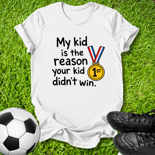 A white graphic t-shirt laid flat on green grass, featuring the text 'My kid is the reason your kid didn't win.' alongside a yellow gold medal illustration.