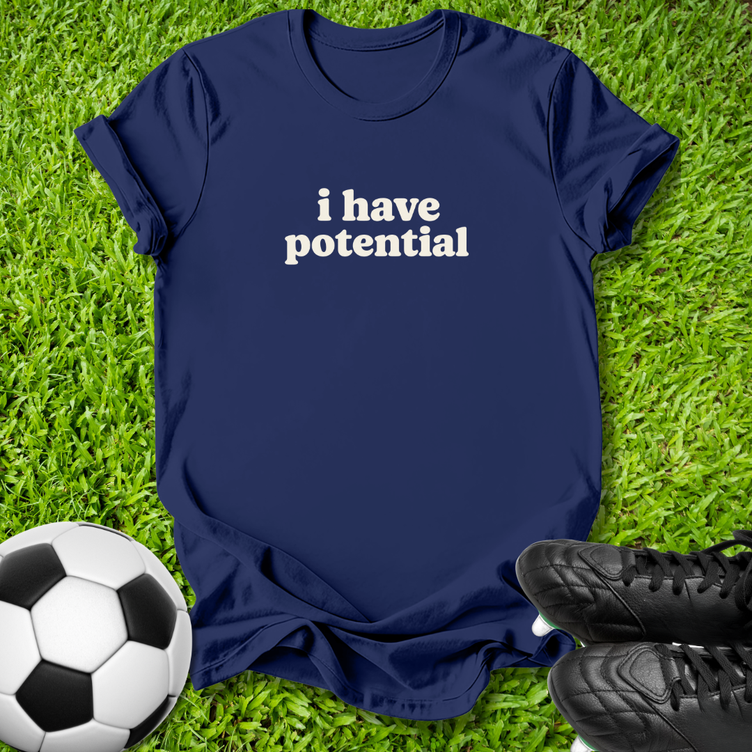 A navy blue short-sleeve t-shirt with the phrase 'i have potential' printed in white across the chest, laid flat on green grass with a soccer ball and black cleats as props.