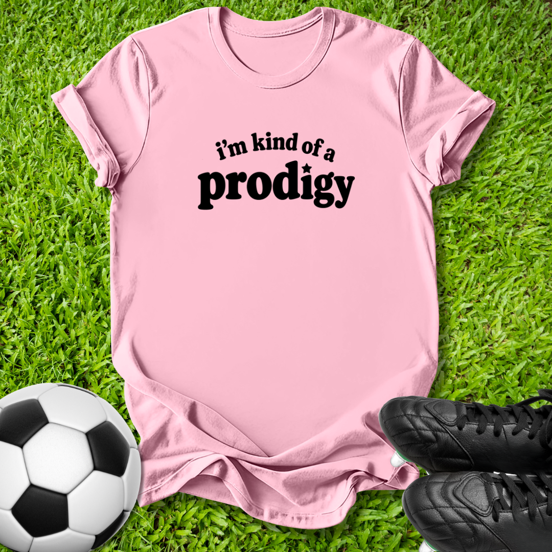 A light pink short-sleeve t-shirt with the black text 'i'm kind of a prodigy' printed on the chest, laid flat on green grass with a soccer ball and black cleats as props.