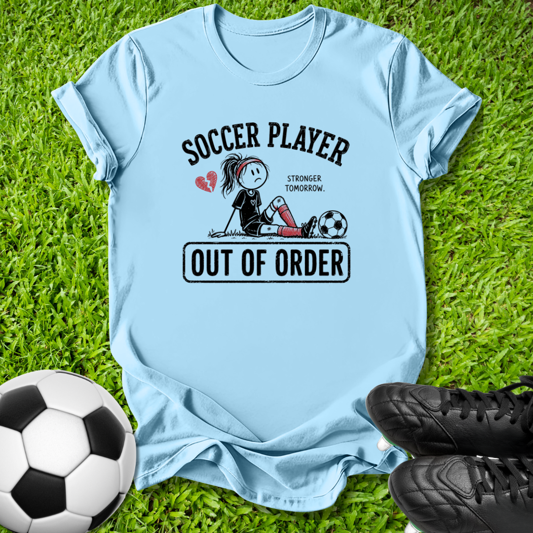 A light blue graphic t-shirt laid flat on green grass, styled with a soccer ball and black cleats. The shirt features a cartoon graphic of a soccer player with the text 'SOCCER PLAYER OUT OF ORDER' and 'STRONGER TOMORROW'.