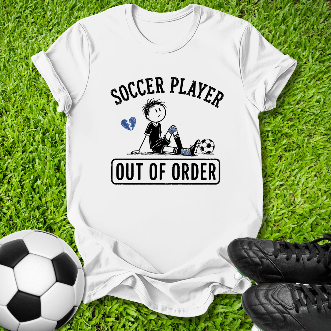 A white graphic t-shirt laid flat on green grass, featuring a cartoon soccer player illustration with the text 'SOCCER PLAYER OUT OF ORDER'. A soccer ball and black cleats are placed around it as props.