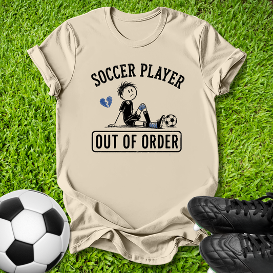 A beige graphic t-shirt laid flat on green grass, featuring a cartoon soccer player design with the text 'SOCCER PLAYER OUT OF ORDER'. A soccer ball and black cleats are placed around it as props.