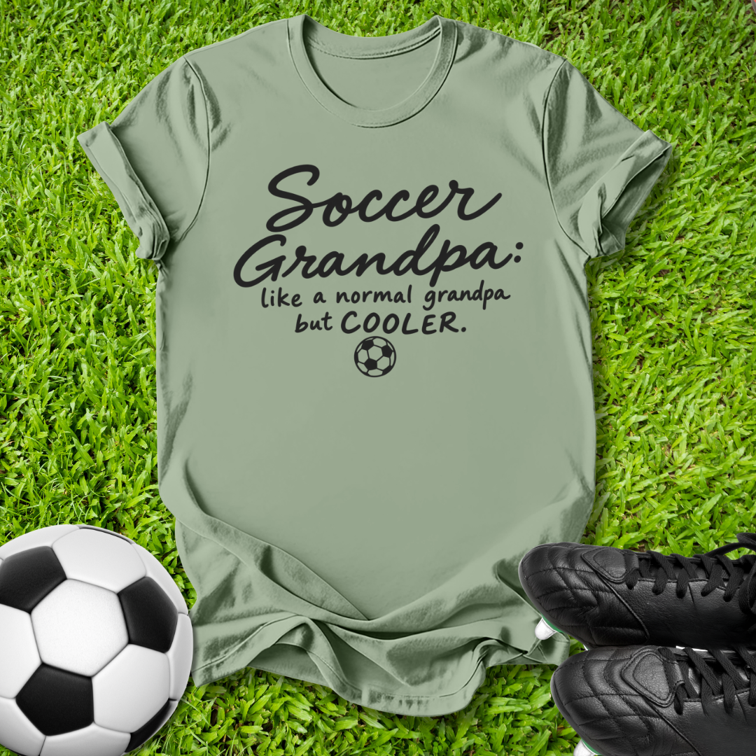 An olive green graphic t-shirt laid flat on green grass, featuring the black text 'Soccer Grandpa: like a normal grandpa but COOLER.' with a small soccer ball icon.