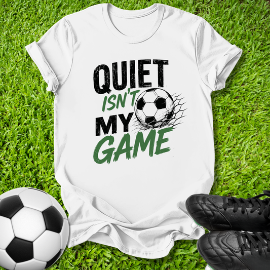 A white graphic t-shirt laid flat on green grass, featuring the text 'QUIET ISN'T MY GAME' in black and green lettering alongside a soccer ball graphic. A real soccer ball and black cleats are placed around it as props.
