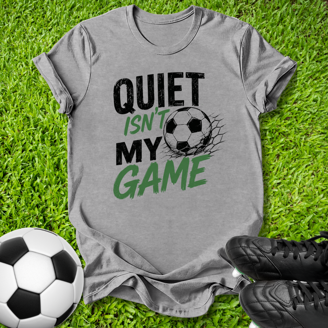 A heather gray graphic t-shirt laid flat on green grass, featuring the text 'QUIET ISN'T MY GAME' in black and green with a soccer ball graphic. A real soccer ball and black cleats are styled around it as props.
