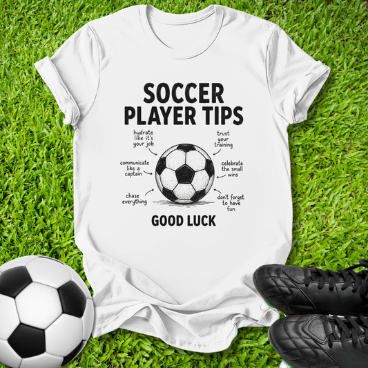 A white graphic t-shirt laid flat on green grass, featuring a black and white soccer ball illustration with the text 'SOCCER PLAYER TIPS' and 'GOOD LUCK' surrounded by handwritten-style tips. A soccer ball and black cleats are placed nearby as props.