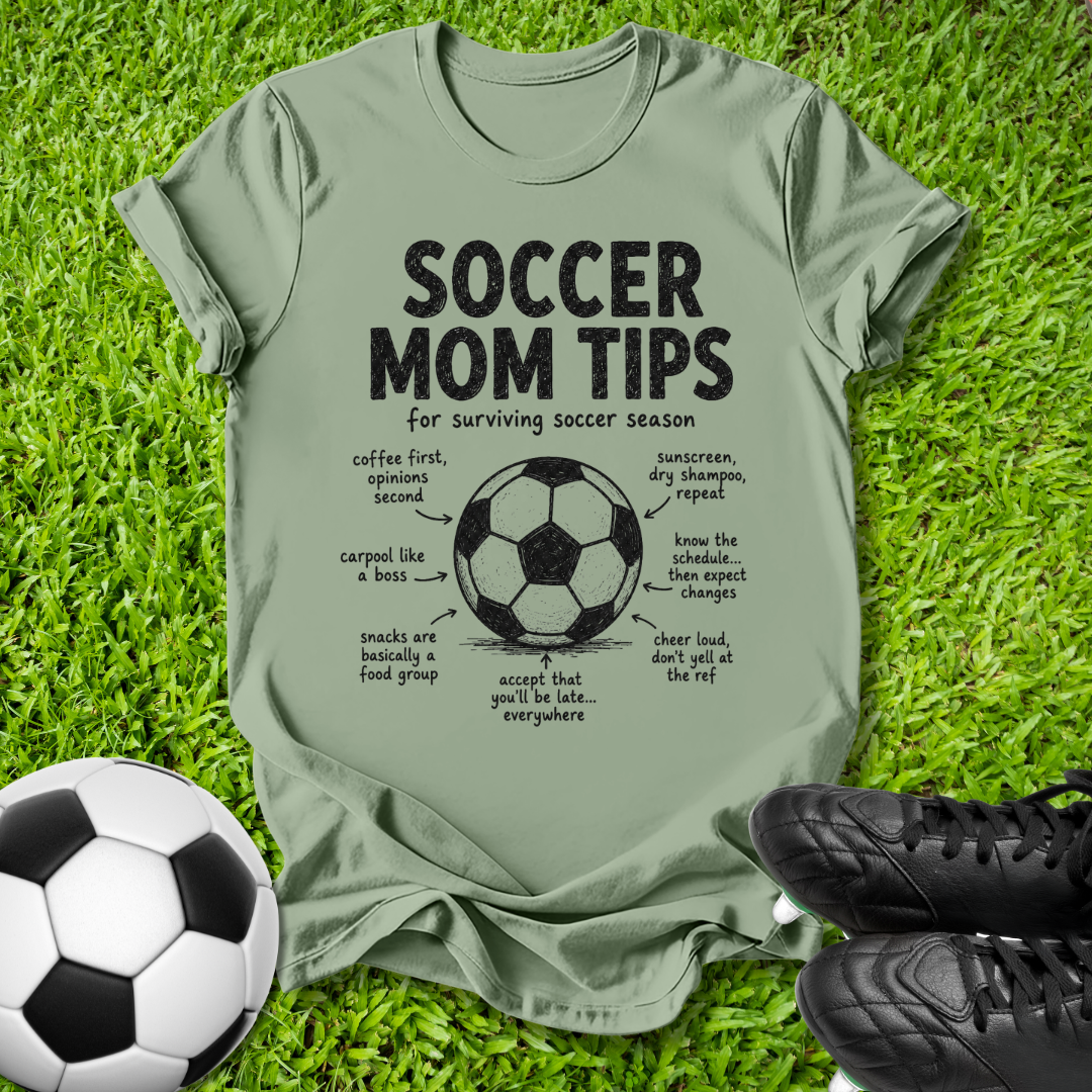 An olive green graphic t-shirt laid flat on green grass, featuring a black and white soccer ball diagram with humorous 'Soccer Mom Tips' written around it. A black-and-white soccer ball and black cleats are placed at the bottom corners of the frame.