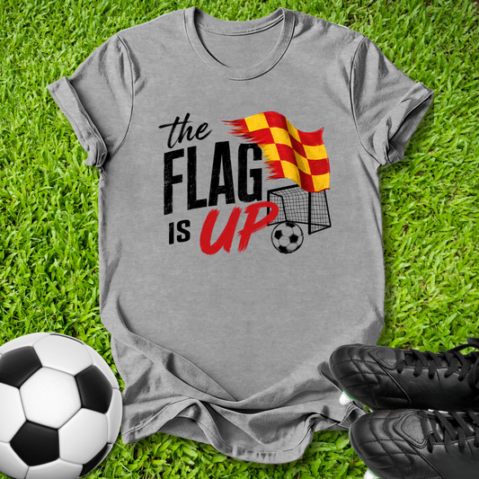 A heather gray graphic t-shirt laid flat on green grass, featuring a soccer theme with the text 'the FLAG IS UP' and a red and yellow striped flag graphic. A soccer ball is on the bottom left and black cleats are on the bottom right.