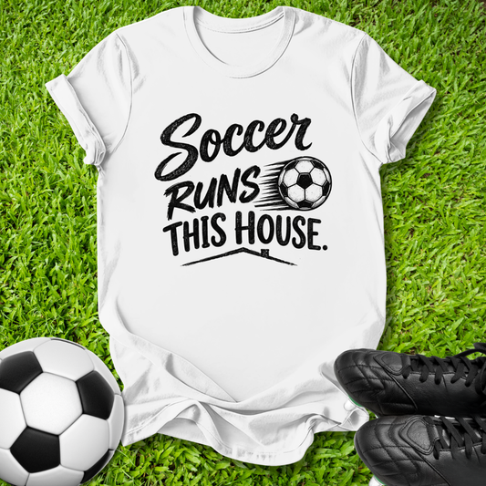 A white short-sleeve graphic T-shirt laid flat on green grass, with a black printed slogan reading 'Soccer Runs This House.' alongside a soccer ball graphic.