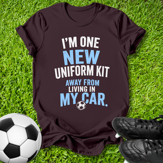 A dark maroon short-sleeve t-shirt with a humorous soccer graphic printed on the front, laid flat on green grass with a soccer ball and black cleats as props.