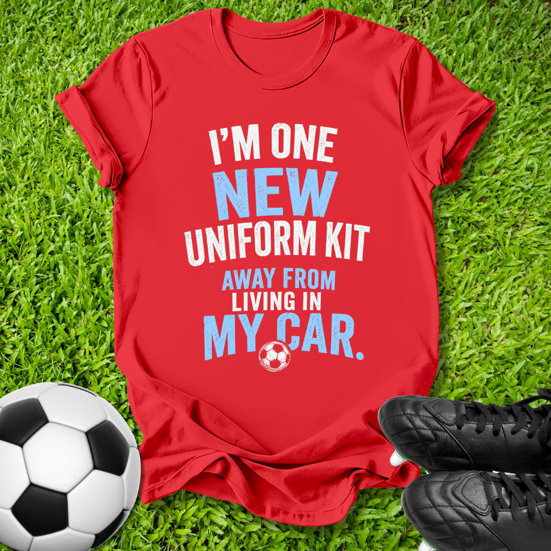 A red graphic t-shirt laid flat on green grass, featuring the text 'I'M ONE NEW UNIFORM KIT AWAY FROM LIVING IN MY CAR.' in white and light blue, with a small soccer ball graphic.
