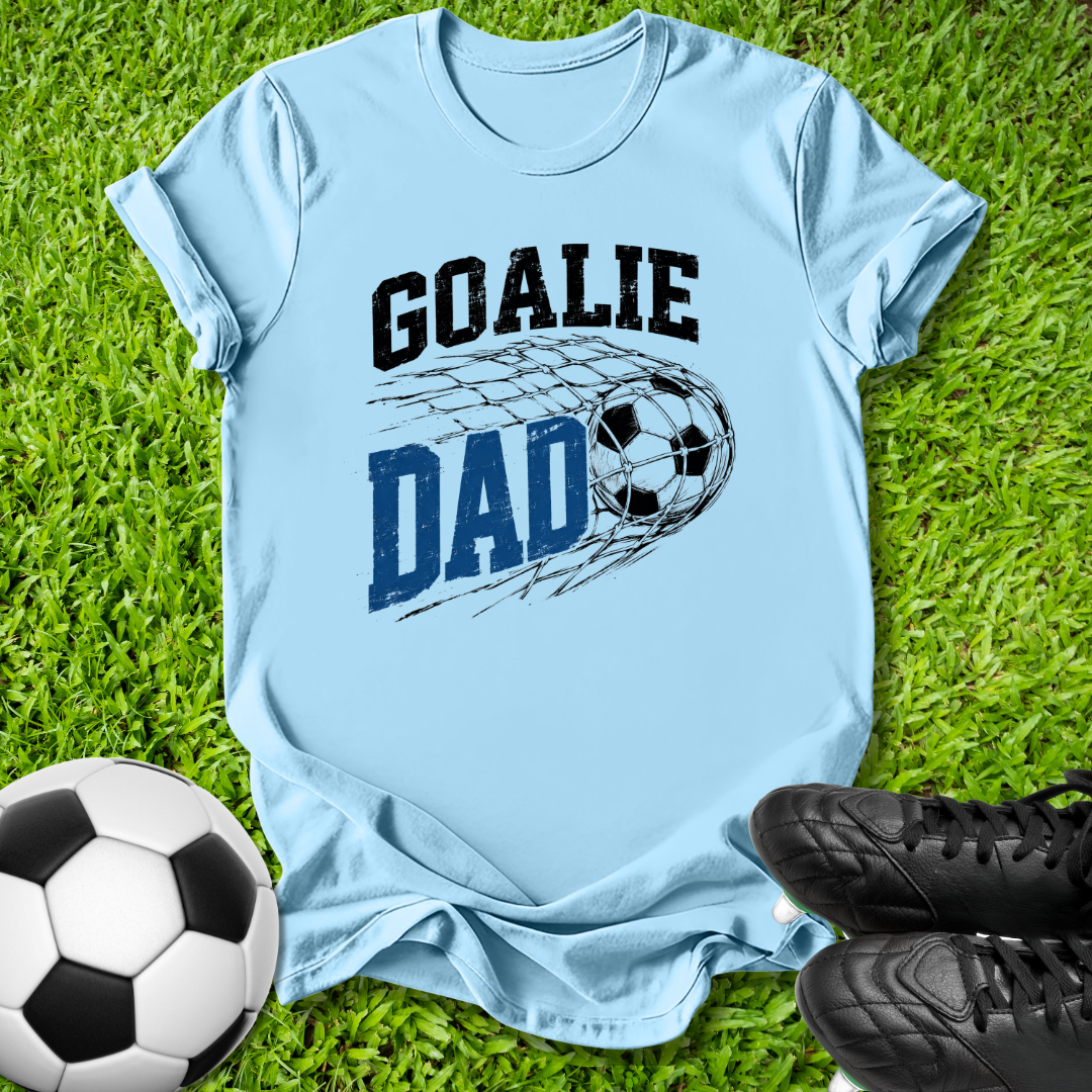A light blue t-shirt with a 'GOALIE DAD' soccer graphic, laid flat on green grass with a soccer ball and black cleats as props.